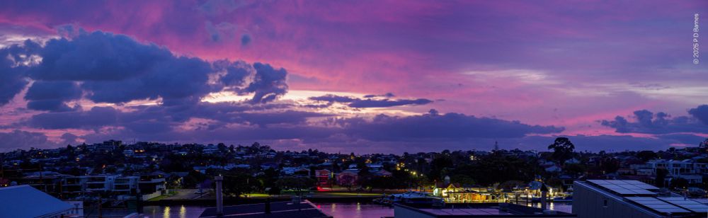 A dramatic cloudscape just before dawn. Two cloud systems provide contrasting colours, shapes and textures across the sky. A high, relatively blank blanket of cloud is lit bright pink, silhouetting lower ragged cumulus which are still dove grey. Light sky can be seen intermittently through both systems, while below street lights and house lights still glow, reflected in the river, and the ferry terminal is a blaze of light.