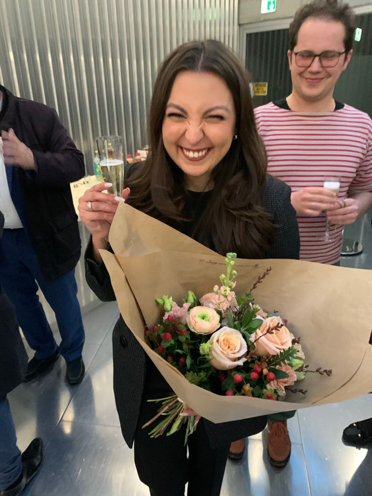 Picture of me celebrating the end of my PhD defence. I’m holding a bouquet of flowers and a glass of Prosecco 