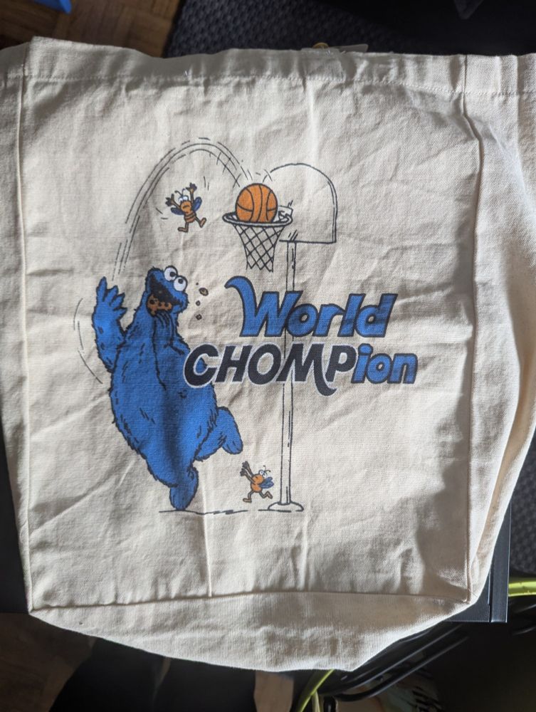 A tote bag featuring The Cookie Monster eating a cookie and throwing a basketball into a net, while two bugs fly around him. The bag features the text World Chompion, as a pun. 