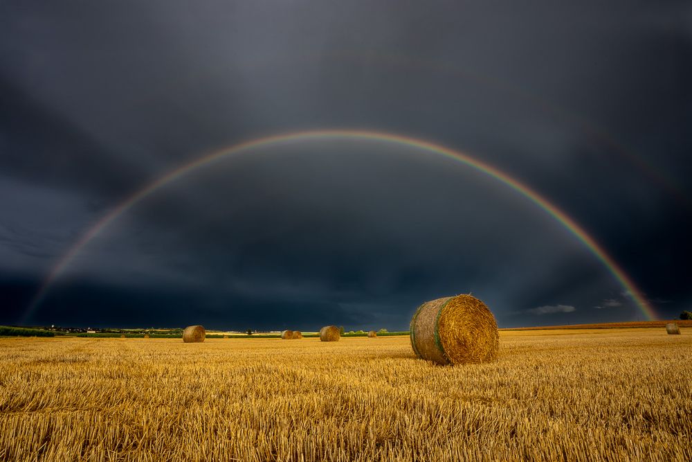 Das Foto zeigt eine weite, goldgelbe Stoppelfeld-Landschaft nach der Ernte. Mehrere große, runde Strohballen liegen verteilt auf dem Feld. Der Himmel darüber ist sehr dunkel und wirkt bedrohlich – fast schwarzblau –, als würde ein Unwetter heranziehen oder gerade abziehen.

Ein strahlend klarer Regenbogen spannt sich in einem weiten Bogen über den gesamten Himmel. Teile eines zweiten, schwächer sichtbaren Regenbogens stehen darüber. Die kräftigen Farben des Regenbogens bilden einen starken Kontrast zu den dunklen Wolken und dem warmen Gelb des Feldes. In der Ferne erkennt man einen schmalen, hellen Streifen Landschaft am Horizont.

Die Stimmung des Bildes ist dramatisch und zugleich friedlich – ein Moment zwischen Sturm und Licht.