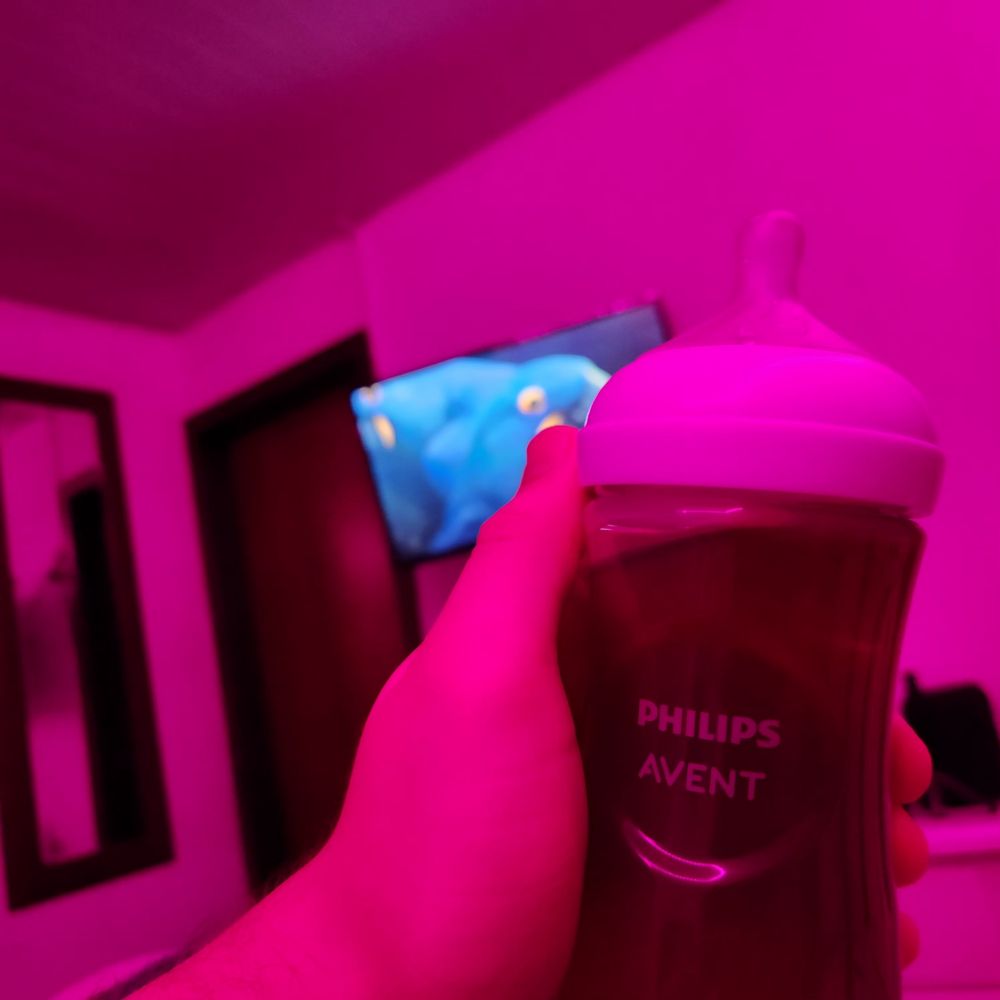 Uma mão segura uma mamadeira Philips Avent em destaque, com um tom rosado-púrpura dominando a cena. Ao fundo, desfocado, há uma tela de televisão exibindo uma arara azul. A iluminação cria uma atmosfera aconchegante e serena no ambiente.