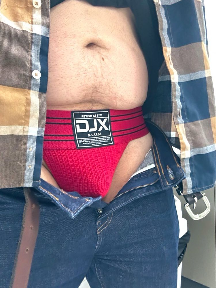 Man in a toilet cubicle wearing a DJX jockstrap in red with jeans and a country style chequered shirt 