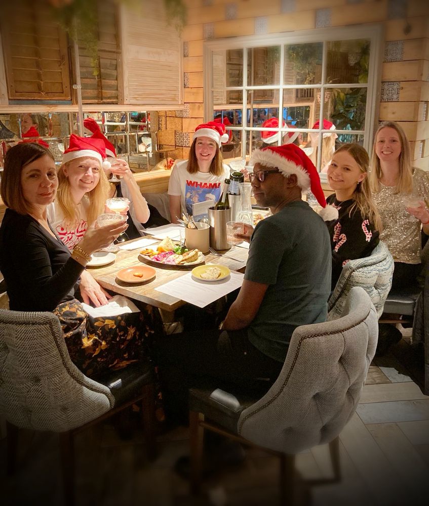 A team of seven work colleagues, seated around a festive table, some in Santa hats and others in sparkly jumpers, raising a toast together! 