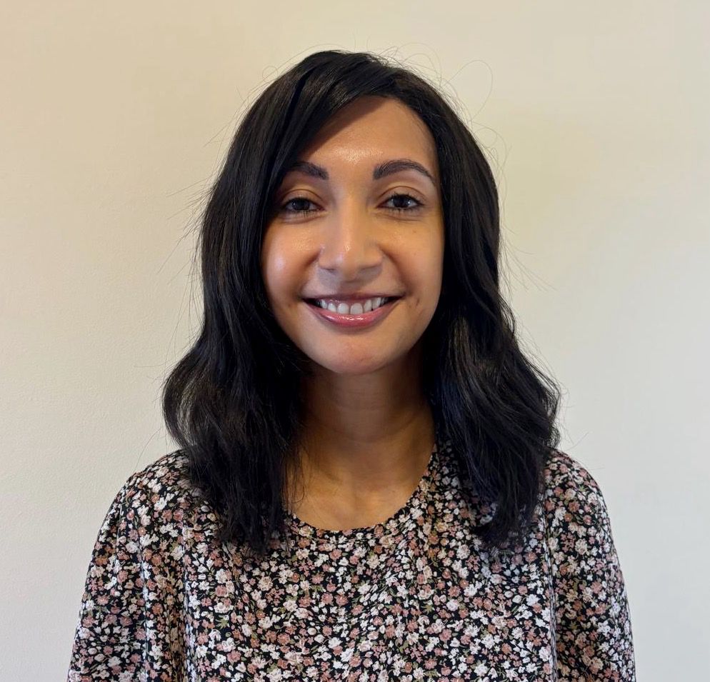 A photo of Saba, smiling and wearing a dark floral top, standing against a cream backdrop