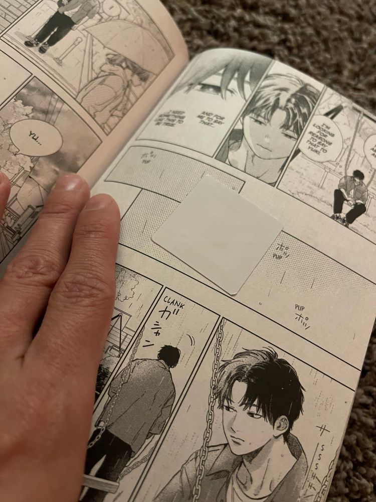 A manga page being held open by my hand. In the middle of the page, there is a security sticker, obscuring some of the drawings in the middle of the manga.