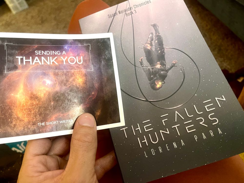Blair holding a thank you card in left hand. The card has a colorful primarily orange galaxy in the back. In my right hand is a book for “the fallen hunters” by Lorena para. It is a white to black gradation galaxy with a person in a space suit upside down. 