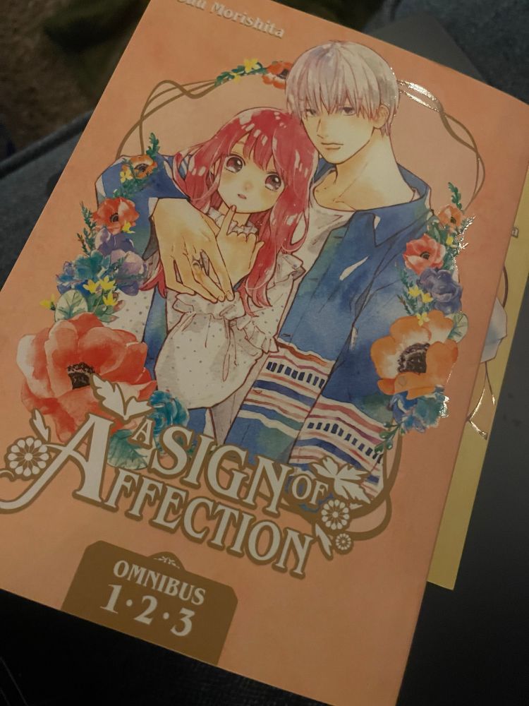 A book cover of “a sign on affection” by suu morishita. The cover is a peach coloring. With two individuals sitting in a frame of flowers. The boy is very tall with shorter white hair. He has his arm around the girl on the left with pink hair. She is holding her hand with her pinky up to her chin. And he is holding that hand also. Their expressions are generally blank. Staring straight that the viewer.