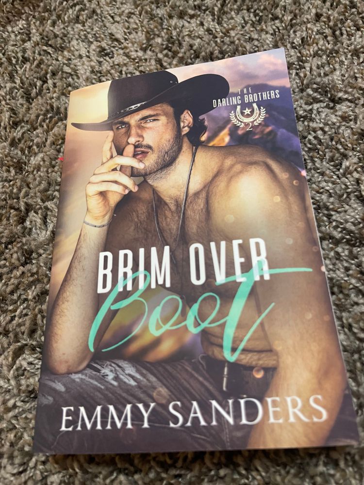 Book cover “brim over boot” by Emmy sanders. Has Montana mountains in the background. And a hunky cowboy posing shirtless with is right hand by his face and other hand hanging loosely 