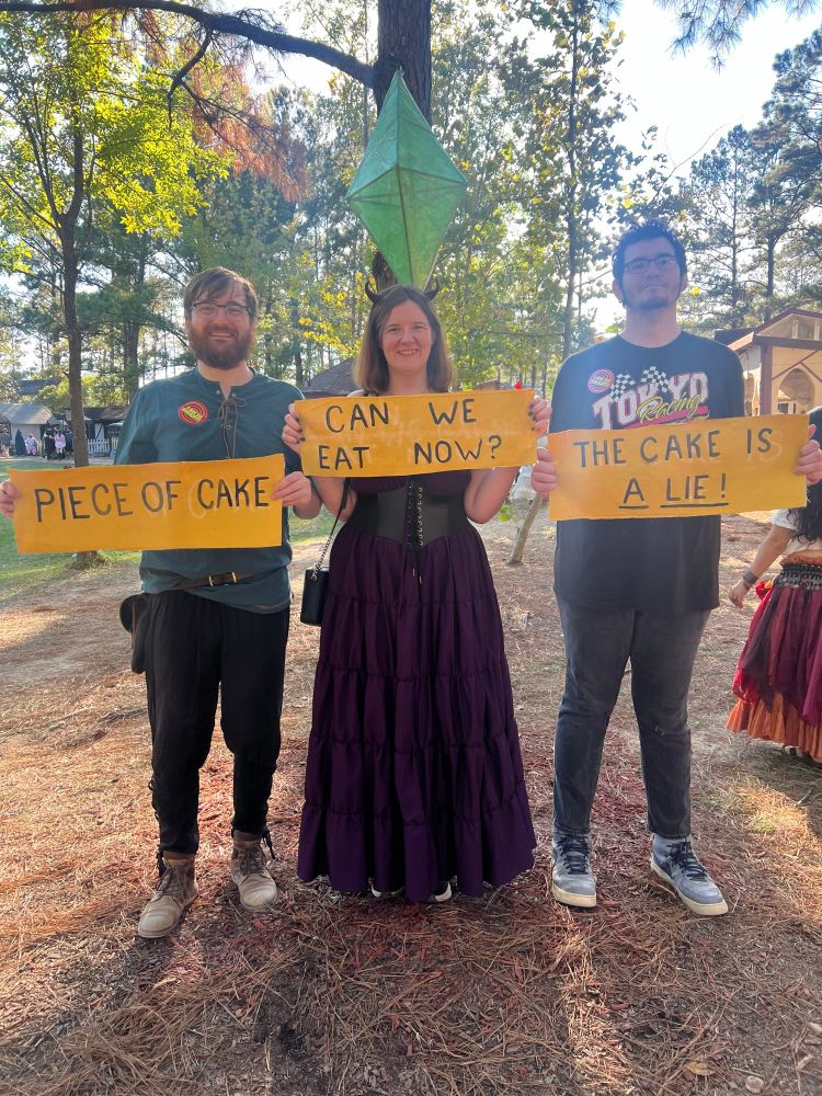 My fiance holding a sign saying piece of cake, me holding ‘can we eat now?’ and my brother holding ‘the cake is a lie!’ A plumbob is above us 