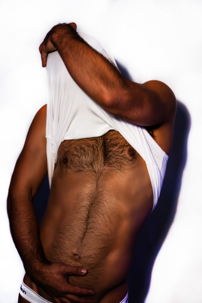 Man holding shirt over his head with his left hand. exposing his hairy chest while his right hand sits on his lower abs. 
