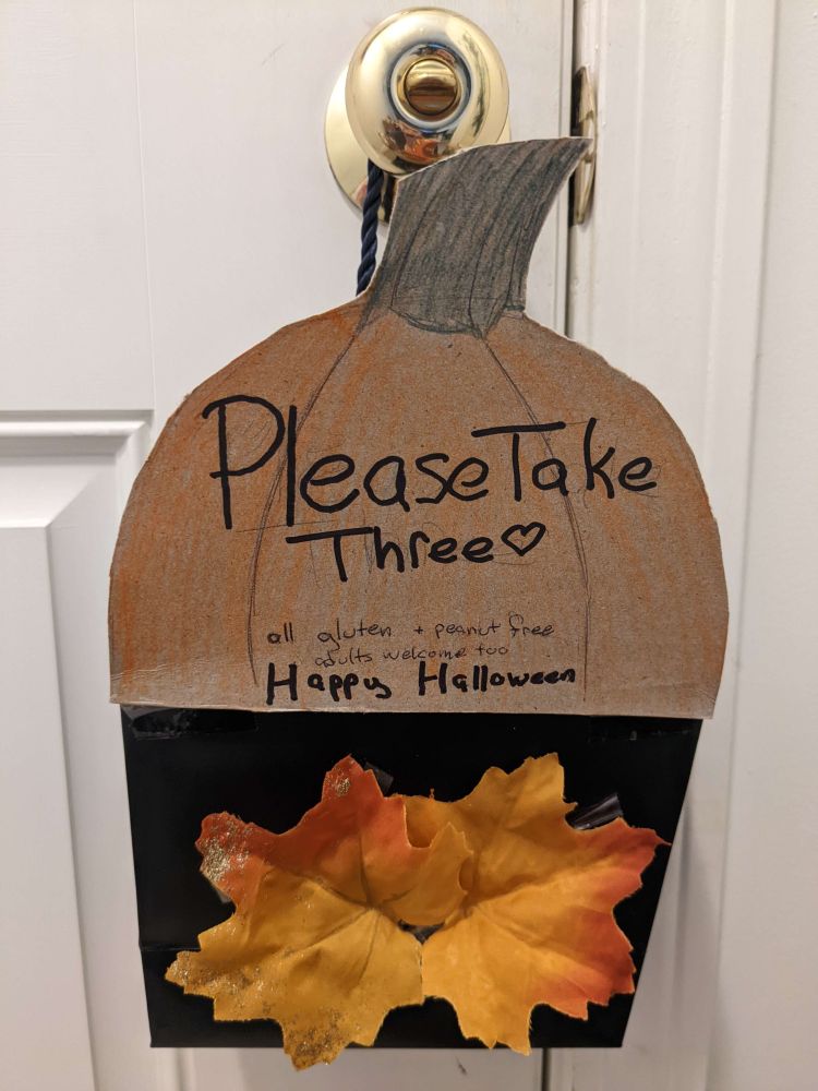 Photo of a Halloween treat bucket made of a stiff paper bag with two orange/gold fabric leaves glued on, and a colored cardboard half-pumpkin on top with directions to "Please Take Three" hung on a doorknob.