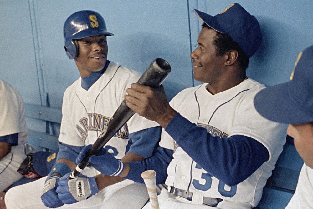 Ken Griffey Jr and Senior on the bench with the Seattle Mariners. They are smiling and holding a baseball bat in the Kingdome dugout. 