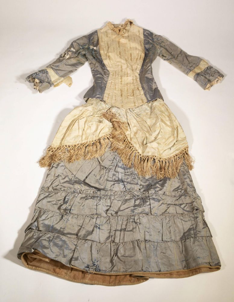 A worn, two-piece Victorian dress laid flat on a white background. The dress features a fitted bodice with long sleeves and a high neckline, made of light blue and cream silk fabric with visible signs of deterioration. The front panel and overskirt are cream-colored with gold fringe trim, while the rest of the garment is light blue with darker blue marbled patterns. The full-length skirt has horizontal gathered tiers and a slight train.