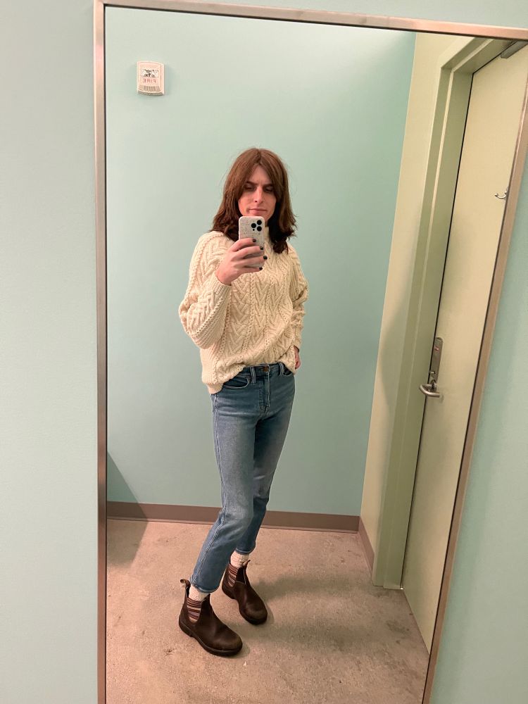 a trans woman is taking a mirror selfie in a changing room. She is wearing a white chunky knit sweater which is French-tucked, high rise straight leg jeans, white socks, and brown blundstones with rainbow stitching