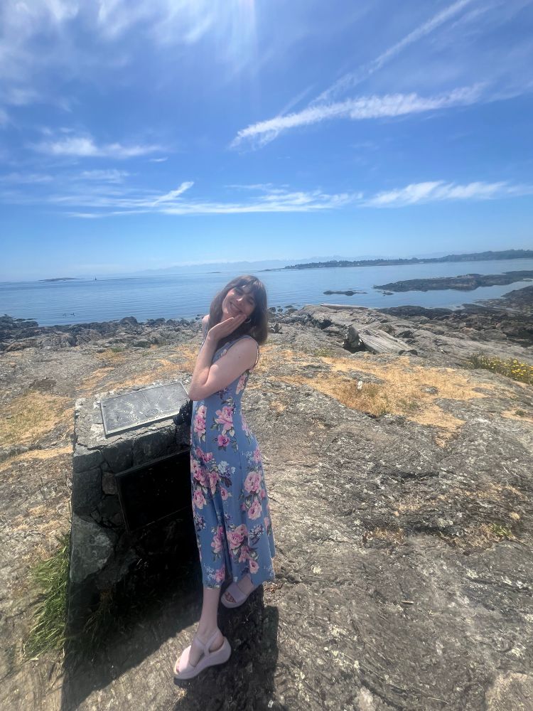 a woman in a blue floral dress wearing lavender platform sandals stands beside a plaque overlooking the sea. Her hand rests on her chin