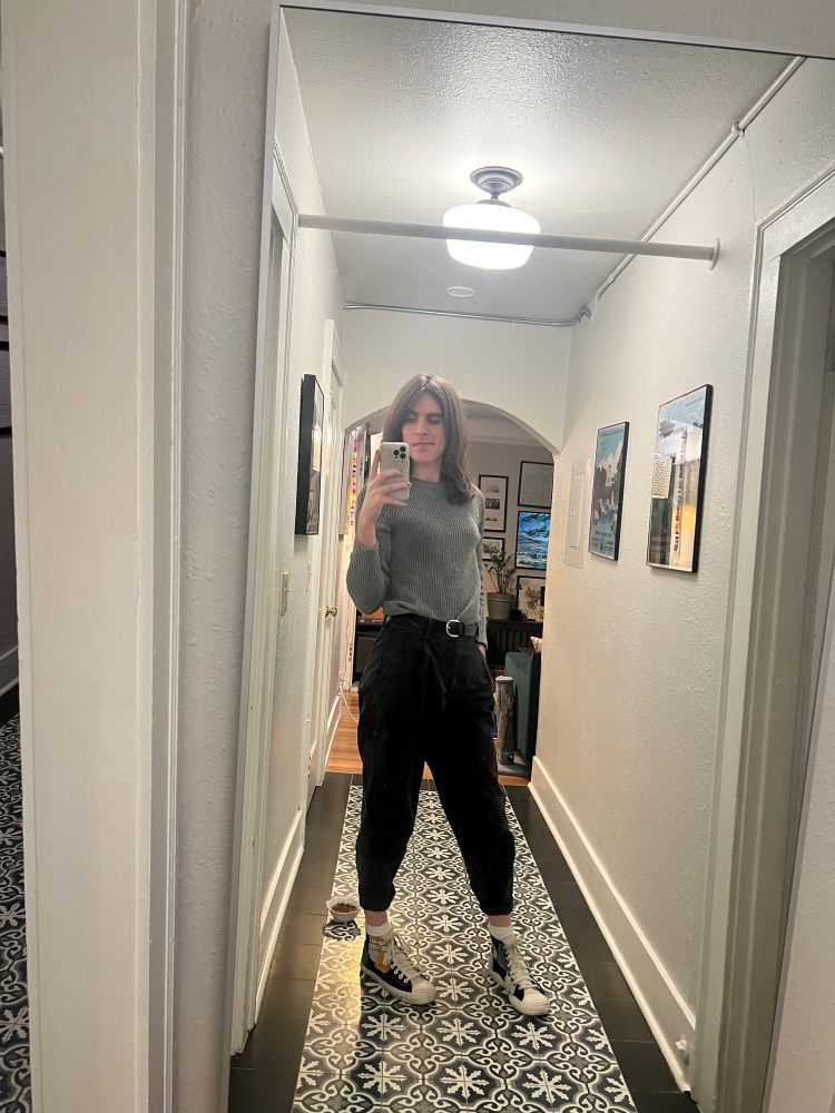 a white trans woman takes a mirror selfie. she is wearing a gray sweater tucked into black pleated chinos with the hem cinched and rolled, and black high-top sneakers with patches of various colors