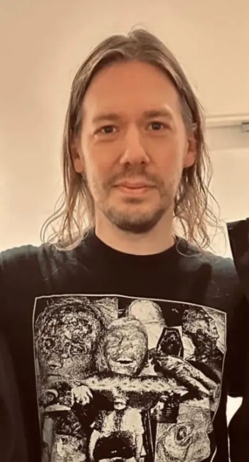 The same man as described in the previous skeet, but standing in front of a blank wall. His facial hair is grown out more but unevenly, and his hair is loose; long enough to brush his shoulders but sort of wavy and scraggly. It kind of looks like it could use a good wash but it's too fine to look nice in this cut and length even if it was clean. 