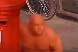 A bald man painted orange hiding behind a post box