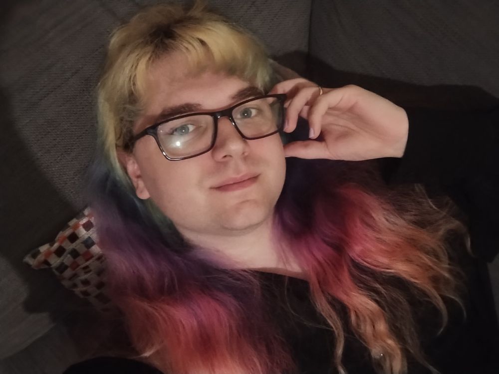 I am lying down on my sofa, my rainbow hair is loose and in front of my shoulders. I am wearing glasses which I am holding with my left hand and looking at the camera.