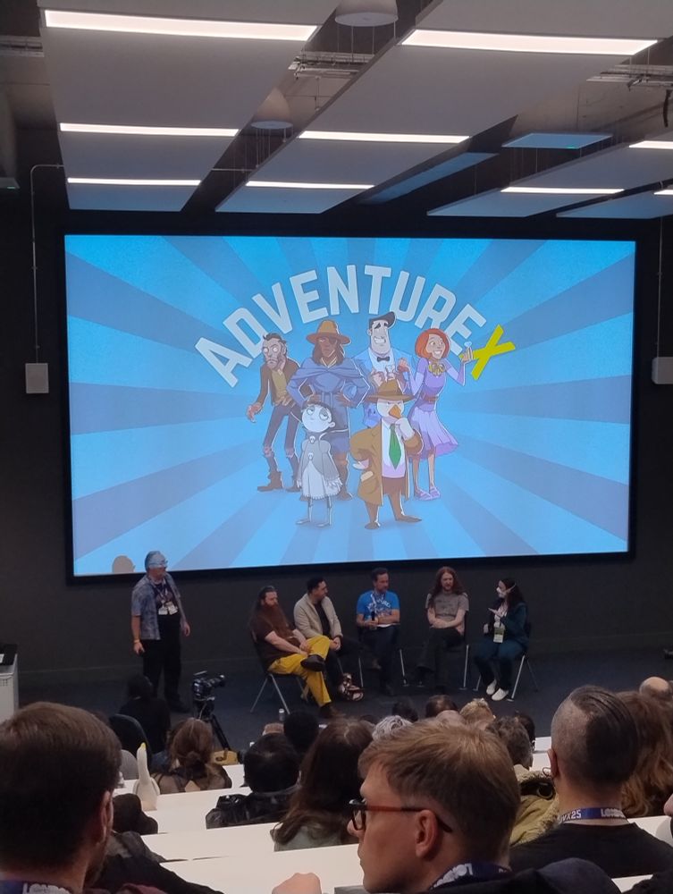 A lecture theatre filled with people. Projected on screen is the AdventureX logo, seated in front are five people in a semi circle.