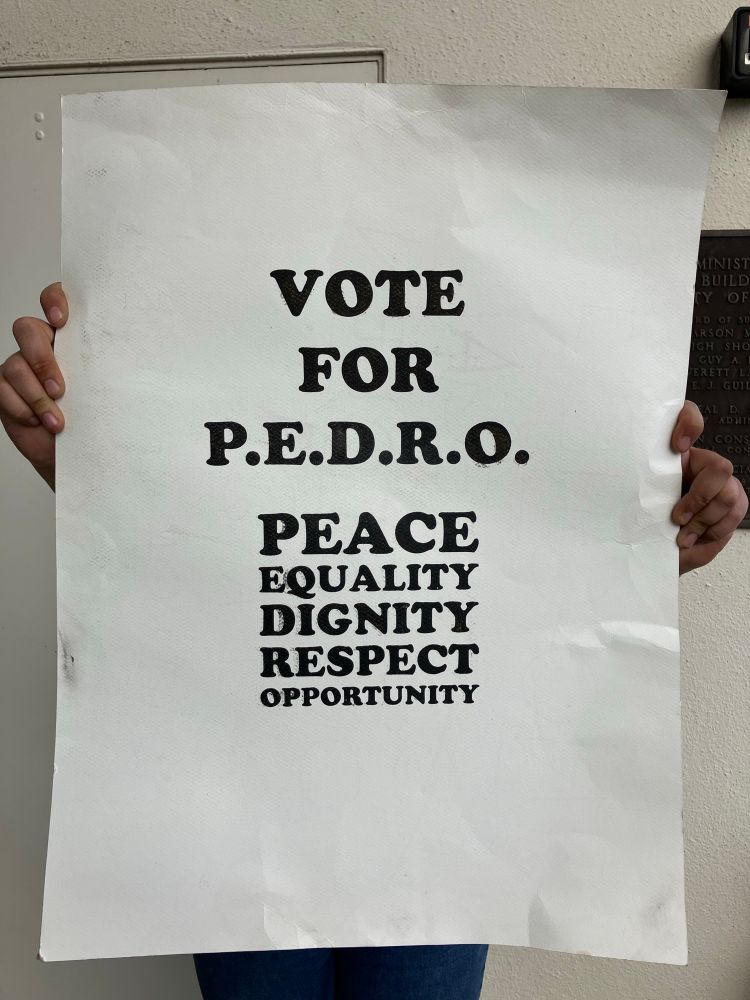 Protestor holding a sign that reads “Vote for PEDRO: peace, equality, dignity, respect, opportunity” (a reference to the “Vote for Pedro” campaign in the film Napoleon Dynamite)