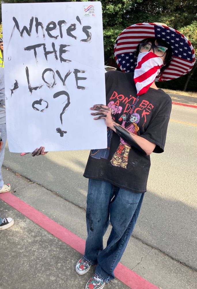 Masked protestor in a US flag hat wearing a shirt that reads “Don’t be a dick,” holding a sign that asks “Where’s the love?”