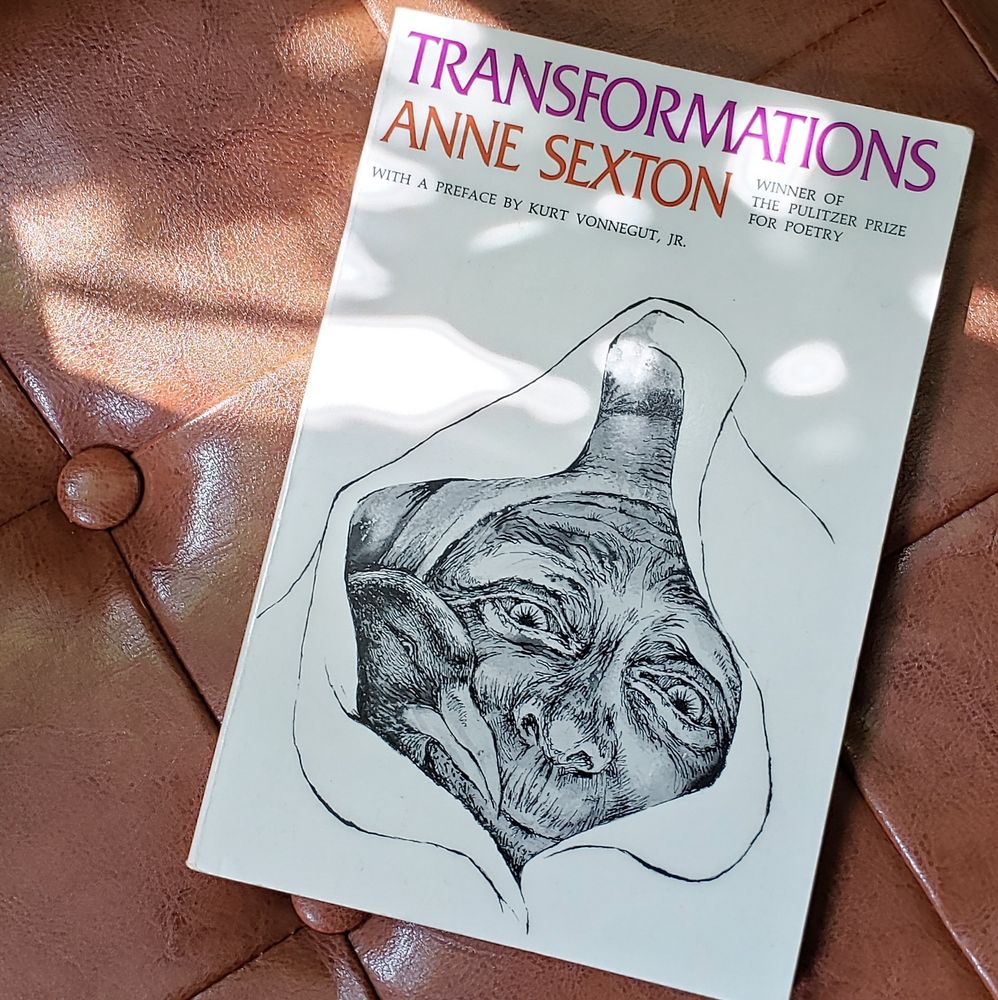 Book lays, cover up, on tan leather footstool. Transformations by Anne Sexton. The cover has an illustration of old woman and a crow peaking through a hole.