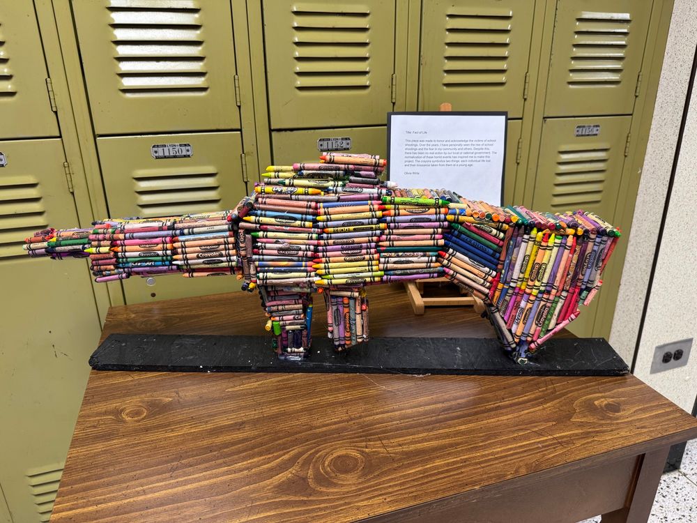 Artist-created “gun” composed of a styrofoam core covered in crayons of different colors. The piece is mounted on a long black board as the base. Behind the artwork are school lockers.