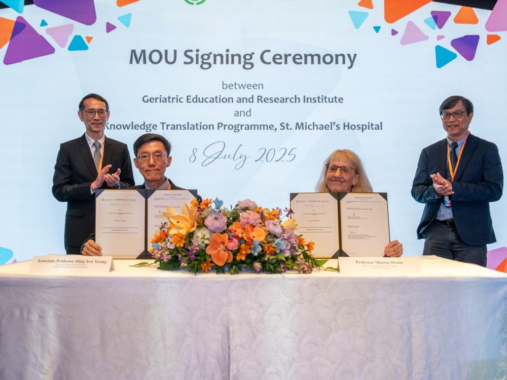 Four people are pictured - two standing and two sitting. The two people sitting are holding up folders with an agreement that they just signed. Behind them, there is a screen that says "MOU Signing Ceremony between Geriatric Education and Research Institute and Knowledge Translation Program, St. Michael's Hospital, July 8 2025". 