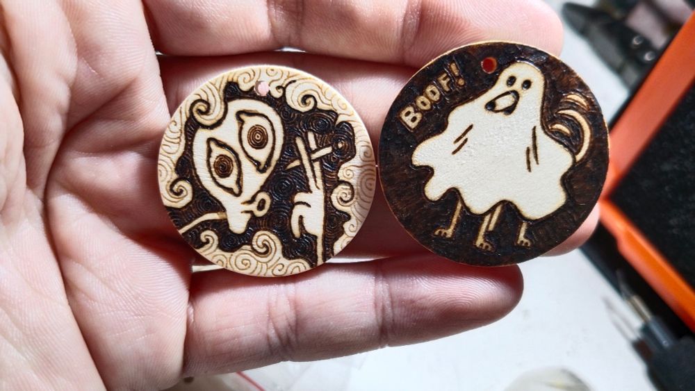 two wooden round cookies, pyrographied. First one has an alien with a blunt surrounded by smoke. Second has a blanket ghost and its dog legs and snoot are showing from under the blanket and mouth hole respectively, they're also waving their tail and saying "boof!"
