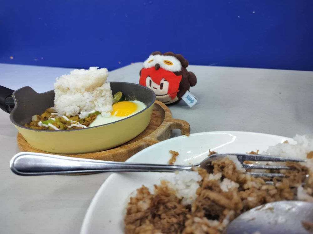 Owluc plushie looking at a mini pan filled with rice egg and sisig. At the right corner is my own plate  with pastil mix into rice cause tangina I ordered a fudging rice meal na sisig instead na sisig lang and punyeta Diluc help me 