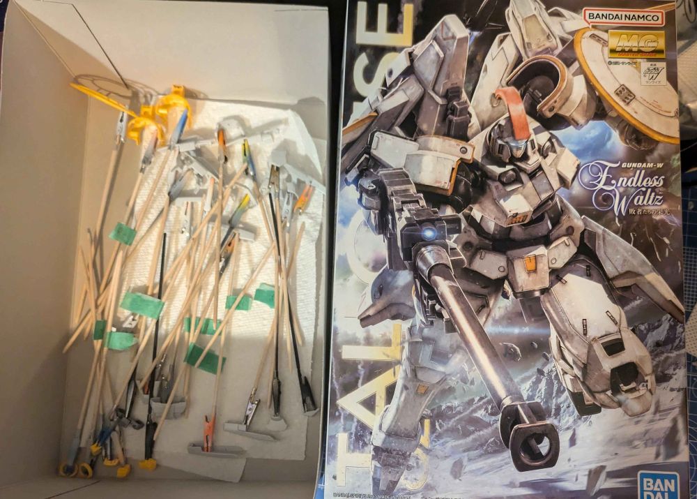 The MG Tallgeese 'Gundam Wing Endless Waltz' gunpla box, and several of its parts set aside on sticks for painting.