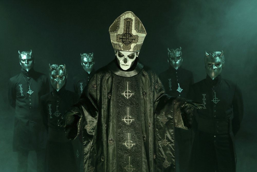 A photo of the band Ghost, their singer bedecked in Bishops mitre and gown, with the Satanic style band logo on both. Behind him, the other five band members stand, wearing black clothes and silver masks with devilish features.