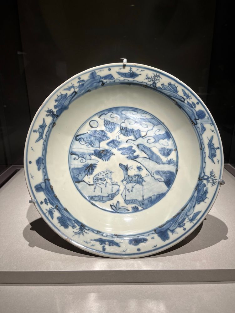 photo of a ceramic blue and white plate. there is art in the center of deer, trees, and clouds and in the lip of the plate of buildings and hills