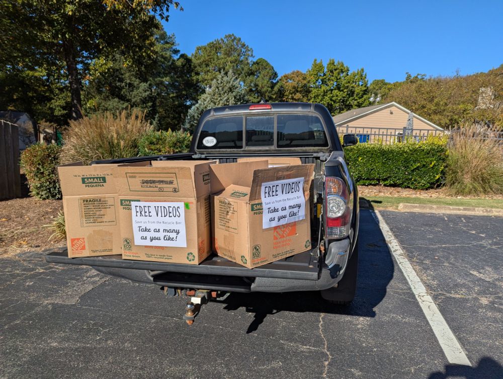 Picture of the back of a pickup truck with cardboard cartons marked "Free Videos, Take as Many as You Like" and "Save us from the recycle bin" printed on them.