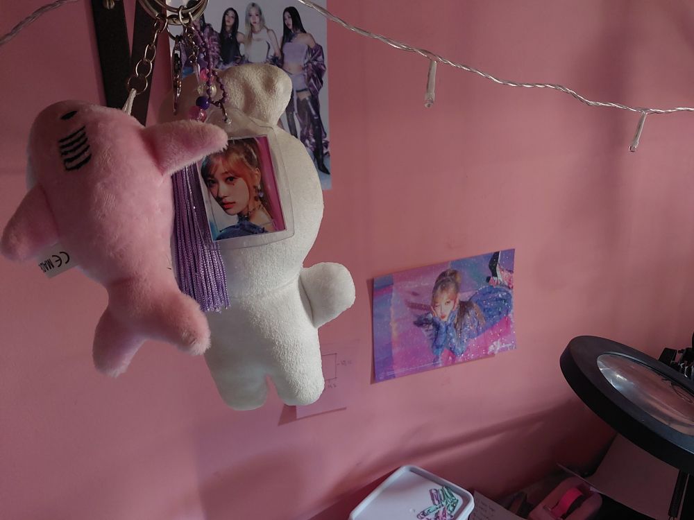 foto de uma parede. não mostra na foto mas tem uma prateleira e pendurados nela tem chaveiros que são visíveis na foto. um deles é uma foto 3x4 da choerry. na parede tem um mini poster a5 da choerry. dá pra ver um do aespa também.