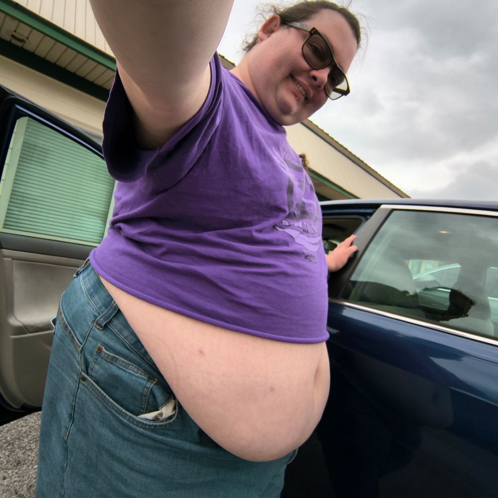 Fat trans woman in the same purple shirt and blue jeans, the shirt has been cropped with a large amount of her belly hanging out over her jeans.