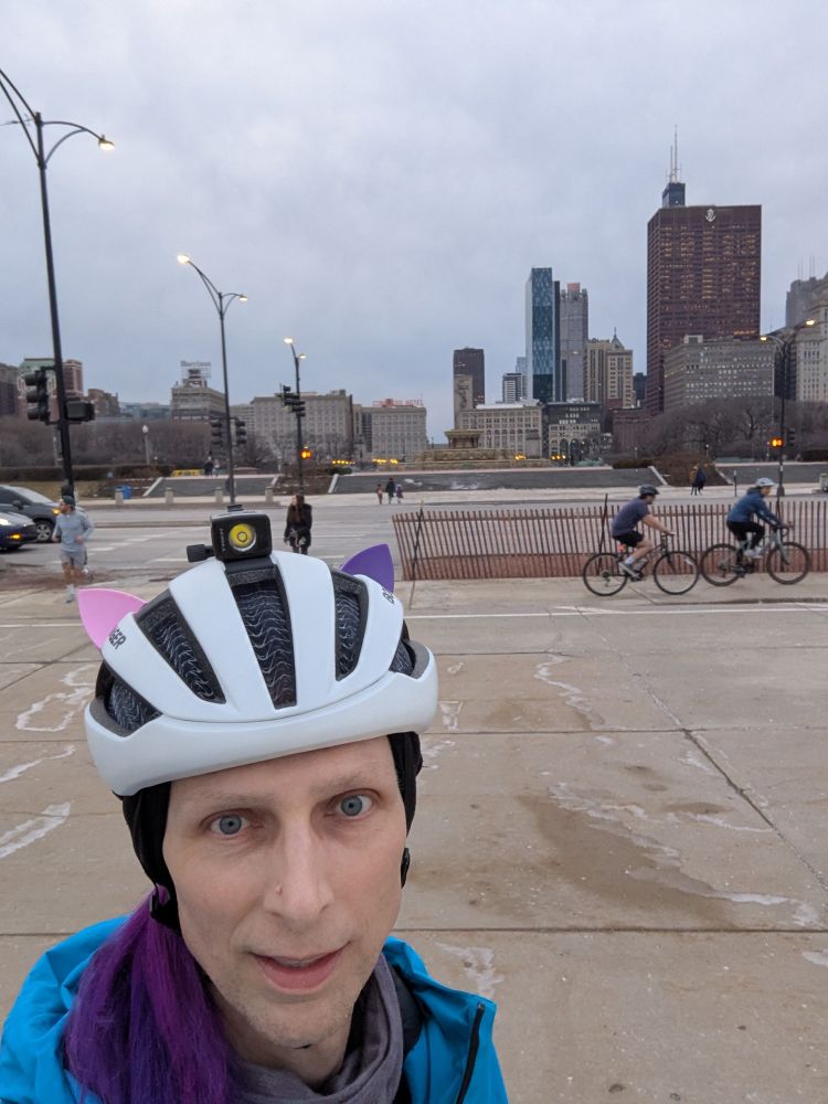 A white woman wearing a white helmet with pink and purple cat ears, and a bright blue coat, takes a selfie of downtown Chicago while 2 cyclist ride in the background 