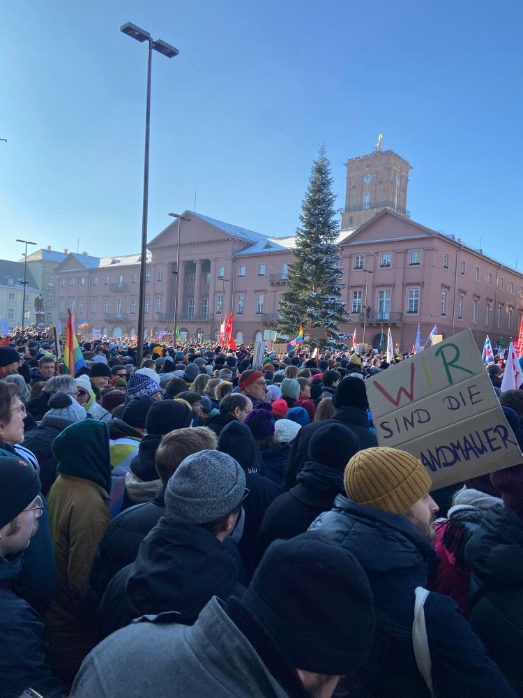 Auf dem Bild ist der Marktplatz in Karlsruhe zu sehen, voll mit dicht gedrängt stehenden Menschen, die für Demokratie und gegen Rechts demonstrieren. Im Vordergrund ein Plakat mit der Aufschrift „wir sind die Brandmauer“.