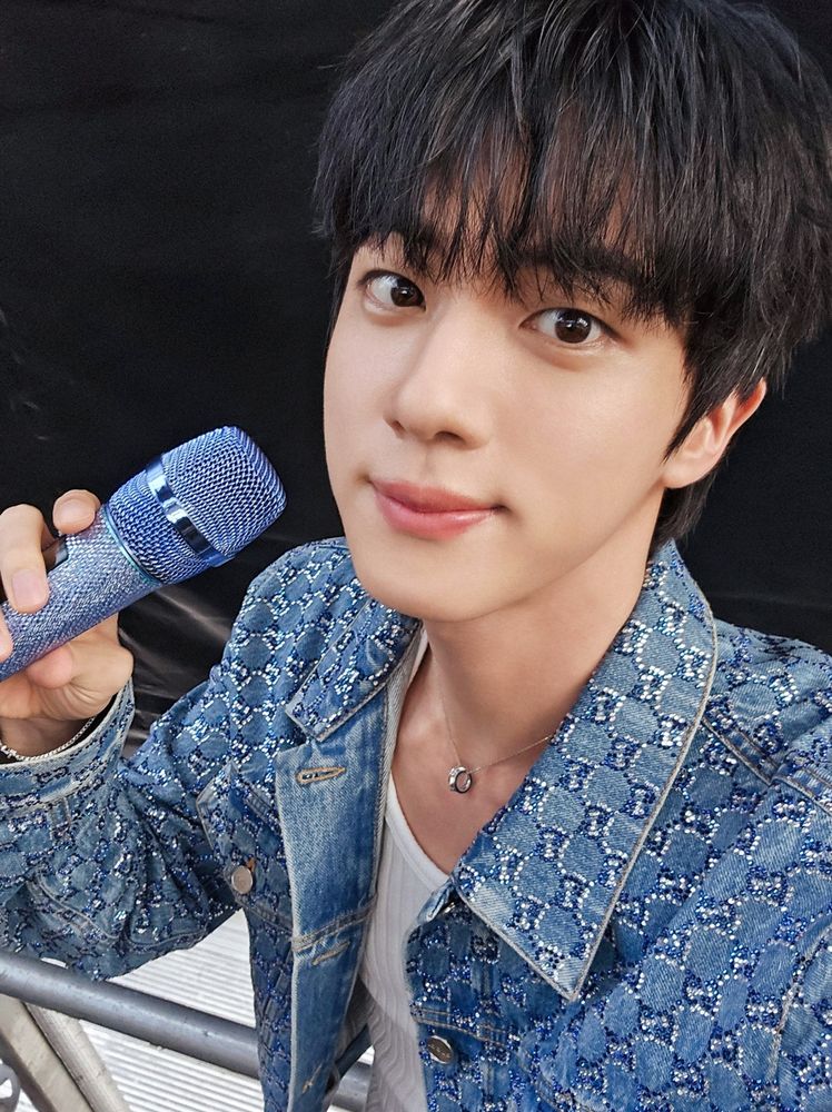 Jin of BTS is holding a blue microphone and wearing a denim, sparkly jacket and white tank top.