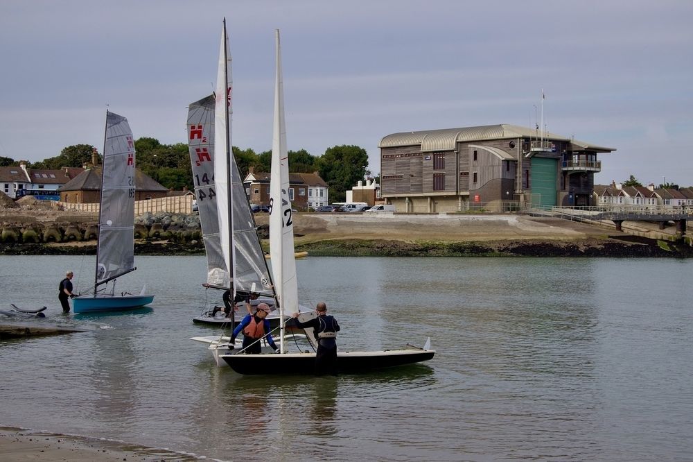 Sailboats with people on board are on a calm body of water near a shoreline with buildings in the background.