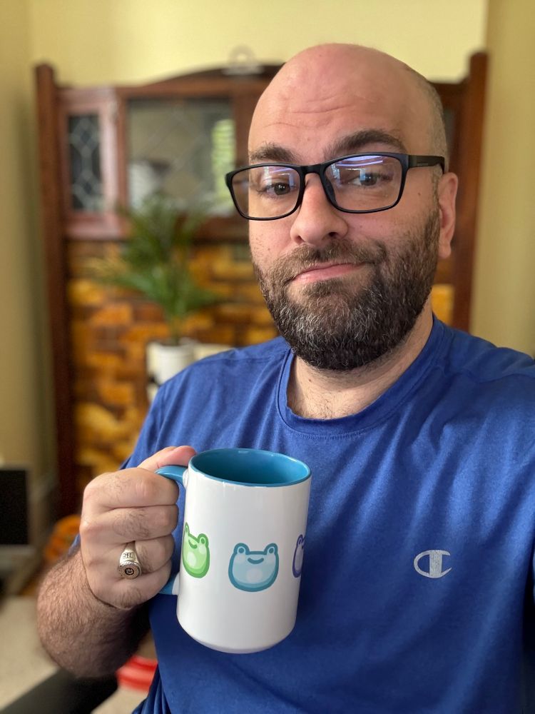 Brian is wearing a blue shirt and holding a white coffee mug decorated with different colored frogs.