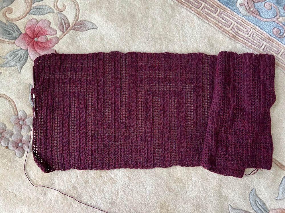 A partially done fillet crochet wrap with a geometric pattern of long way stripes overlaid with alternating bars that go 3/4 of the way across