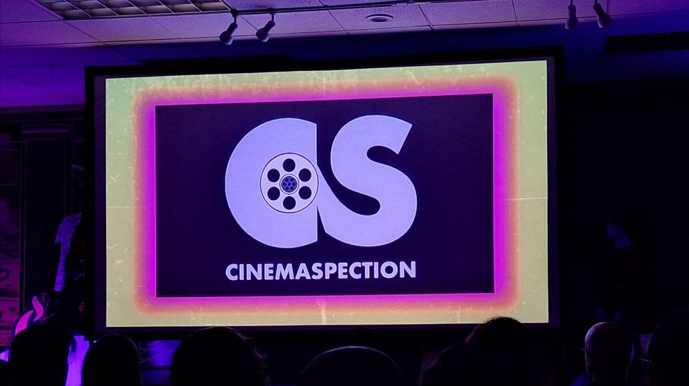 The logo for CinemaSpection, consisting of a stylized "C" and "S" with a film reek nestled in the c, being projected on a big screen in front of an audience.