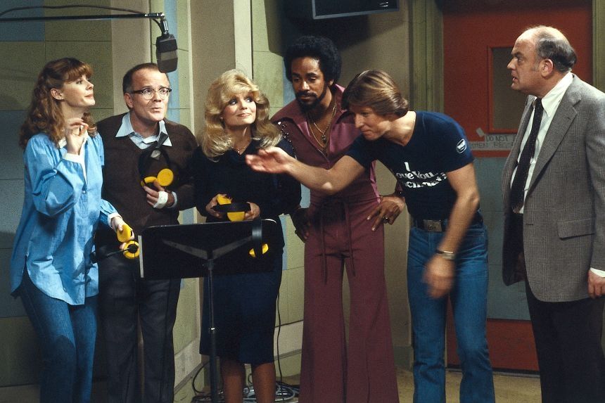 An image from WKRP in Cincinnati showing Jan Smithers, Richard Sanders, Lori Anderson, Tim Reid, and Gary Sandy all standing under an overhead microphone while Gordon Jump looks on.