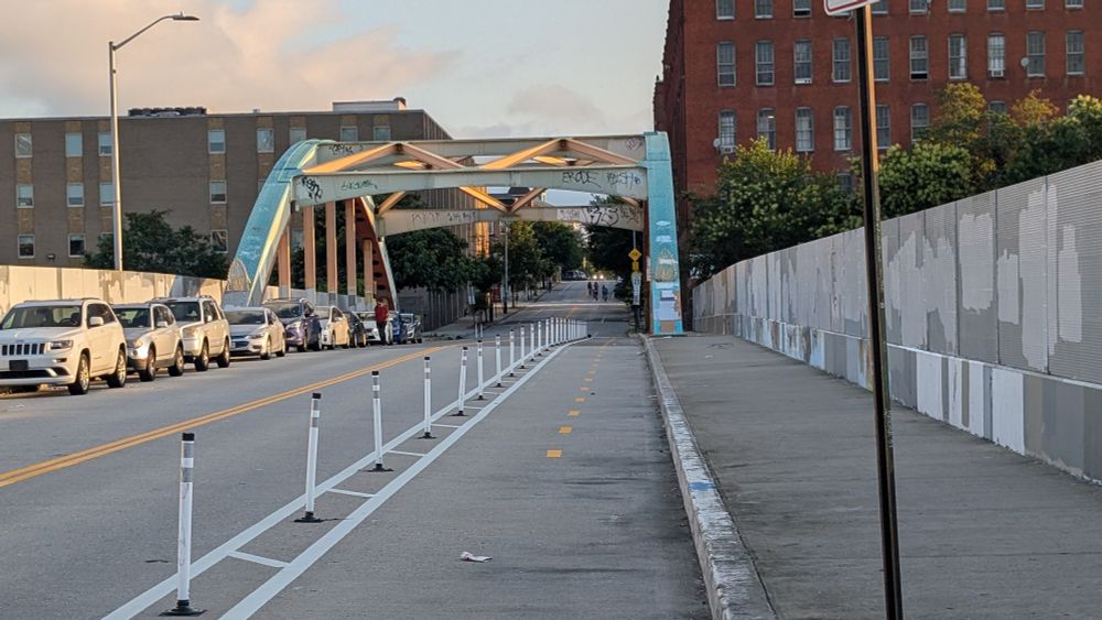 A two-way, flex post divided bikeway on an urban street with a steel frame bridge in the background in the soft morning light. 
