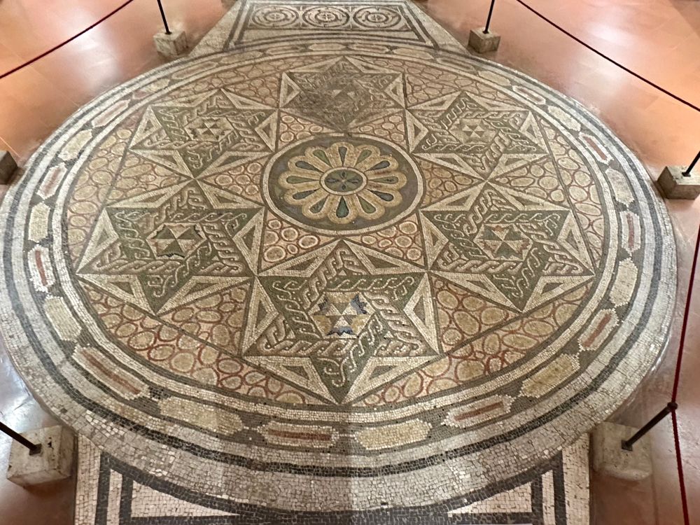 A circular floor mosaic, with a “wheel” design in the centre. Diamond shaped patterns surrounding, formed of inverted triangles with interlace borders.