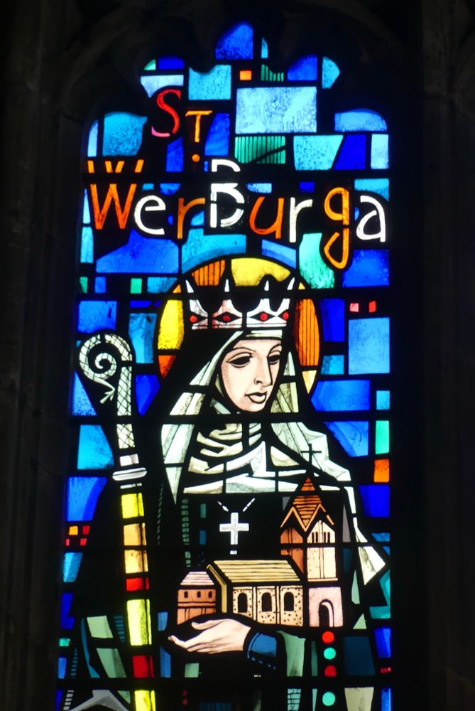 A bright background of different blue glass. The Anglo Saxon princess is wearing a crown, but with a halo. She is dressed as a nun, holding a crozier & a model of the church.