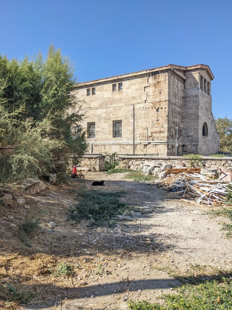 A black cat from afar lies in the middle of a gravel road with a stack of construction materials off to its right and a stone building in the background.
