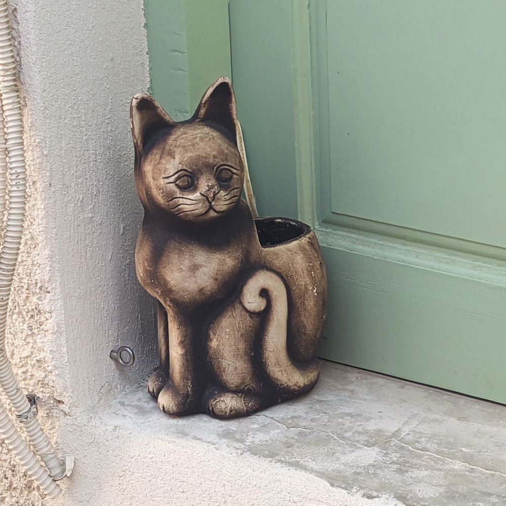 Not a real cat: a plant vase shaped like a cat with a tail curled up to its chest stands on the stoop of a green door in a village in naxos.
